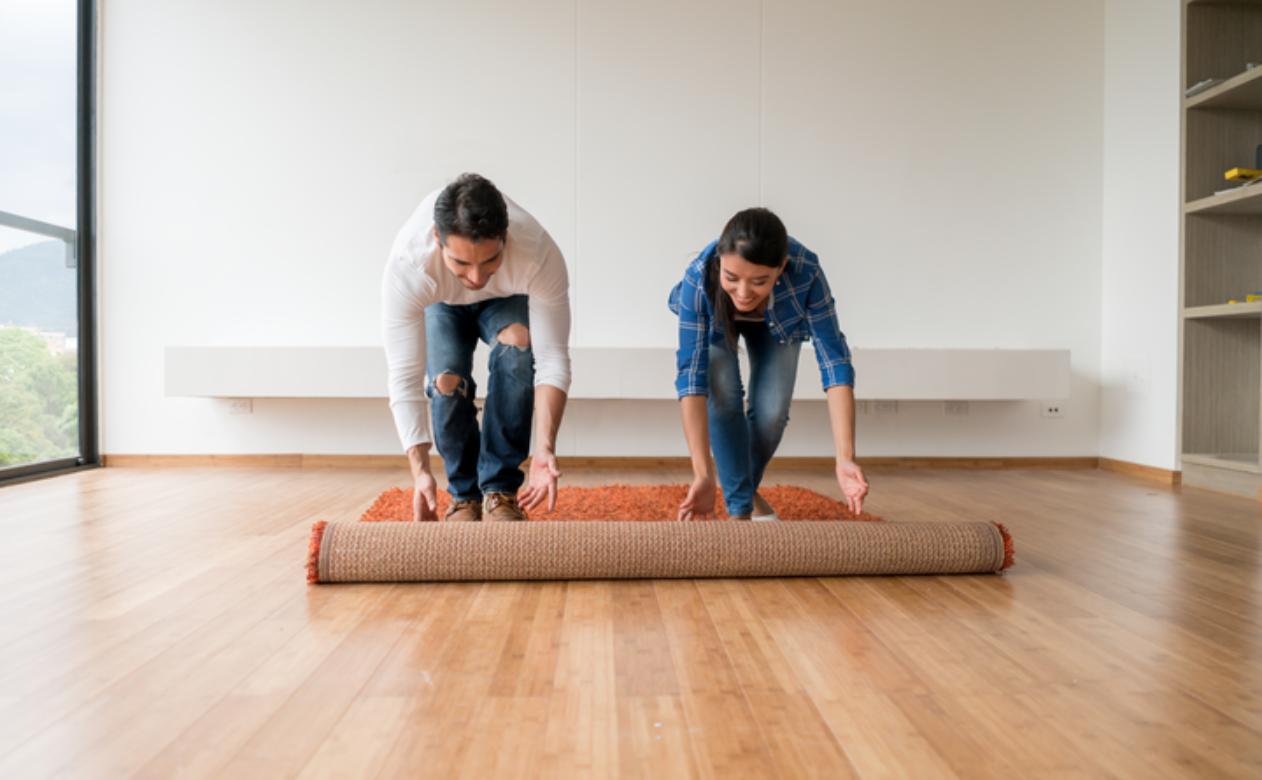 two people unrolling orange carpet in room with wood looking flooring and large windows.